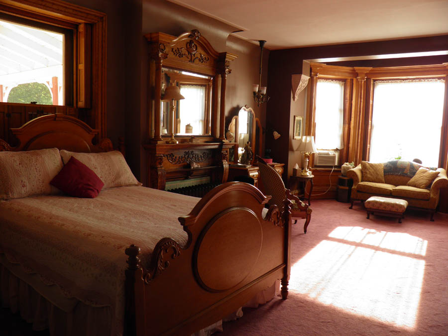 Victorian-style bedroom with an ornate wooden bed and dresser, a sunlit sitting area with a sofa in the background, in warm brown and cream linocut tones.