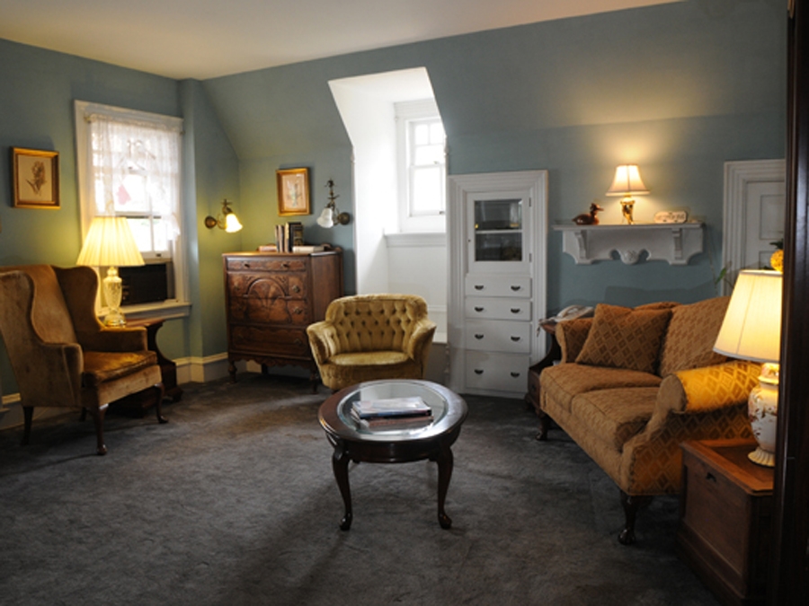 Carved linocut relief print of a sitting room with antique armchairs and sofa, wooden dresser and cabinet, soft lamp lighting, muted blue-green walls, warm cream and sage tones